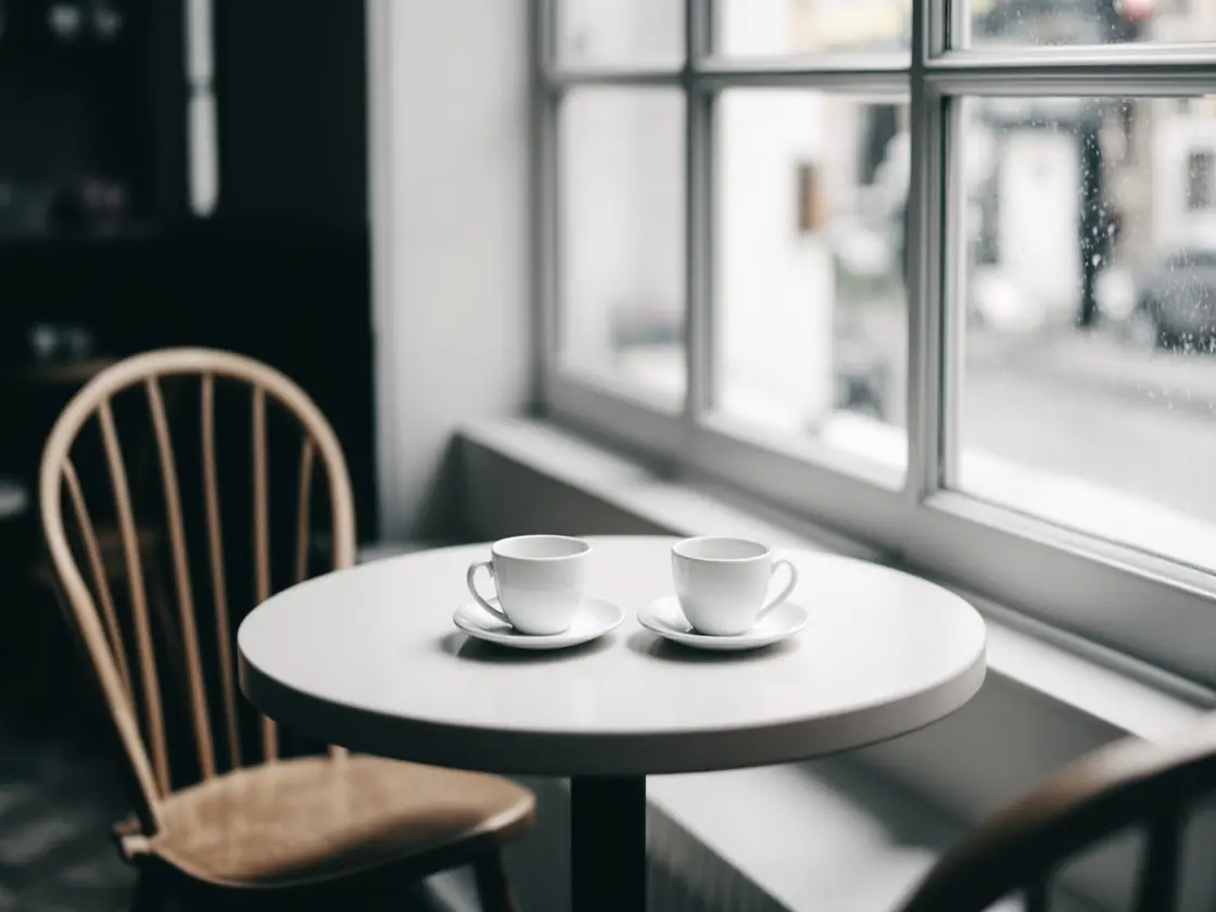 A small round cafe table with two coffee cups beside a window