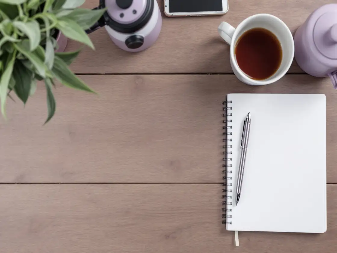 A notebook, a pen and a cup of tea on a wooden desk seen from above